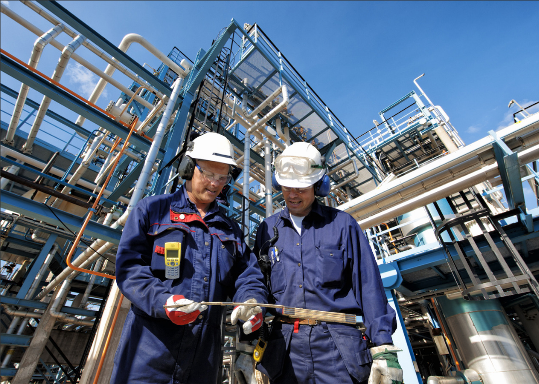 Two men in blue jumpsuits and helmets having a discussion at a petroleum plant.