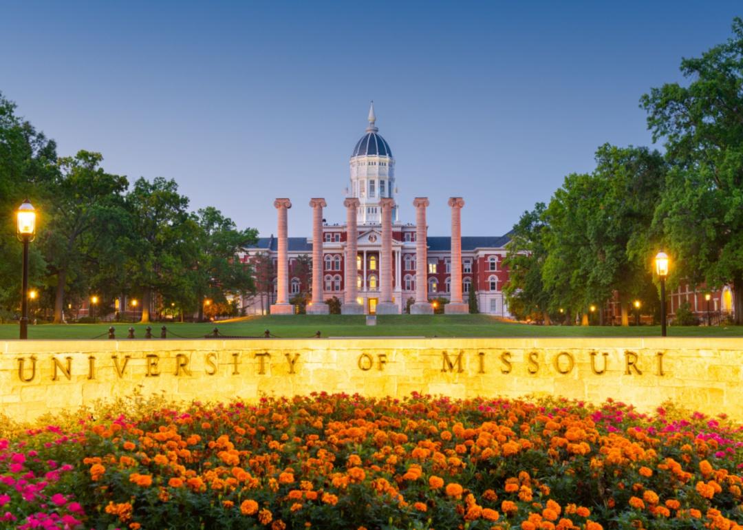 A large historic brick builing with large columns on the lawn in front and a lit stone sign and flowers in the foreground. 