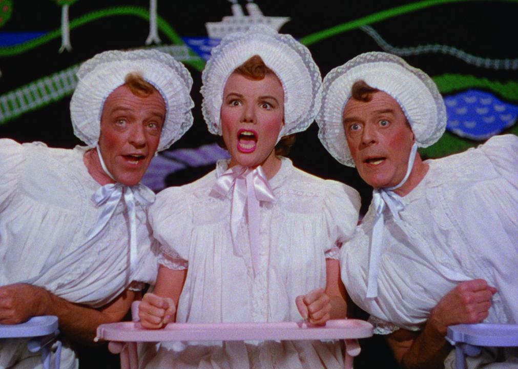 3 adults dressed in white baby gowns and bonnets sit in high chairs.