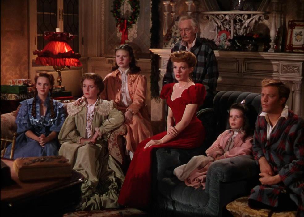 A family sits in the living room in their nightclothes with Judy Garland in a red gown in the center.