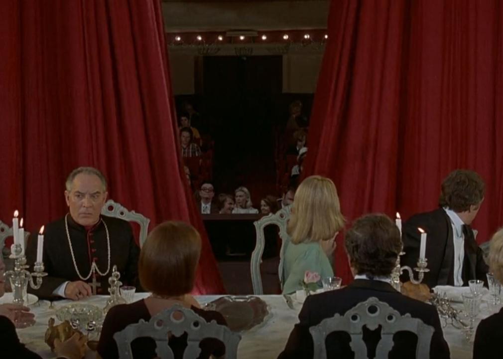A group of people sit at a dinner table while a red theater curtain opens around them revealing an audience.