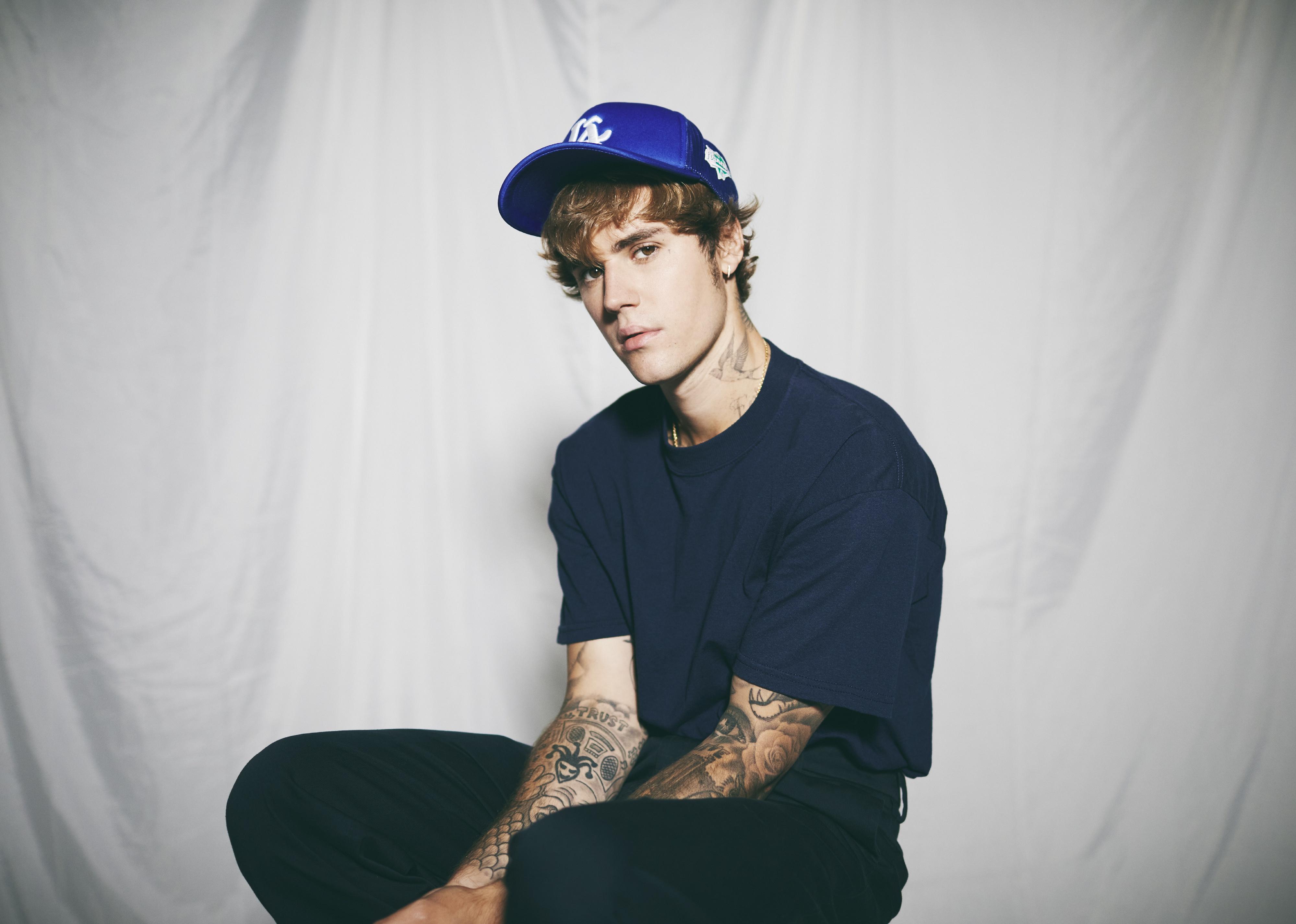Justin Bieber sitting in a studio wearing a blue baseball cap, blue t-shirt and pants.