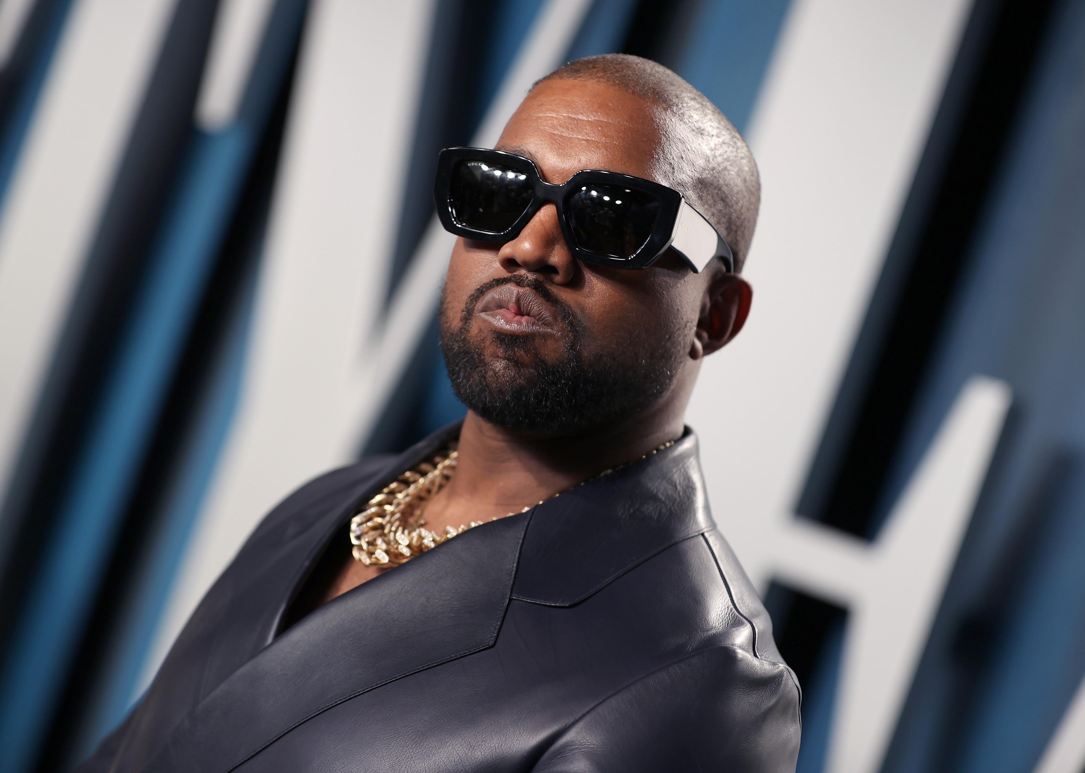 Kanye West in a black leather jacket, gold chain and large dark sunglasses.