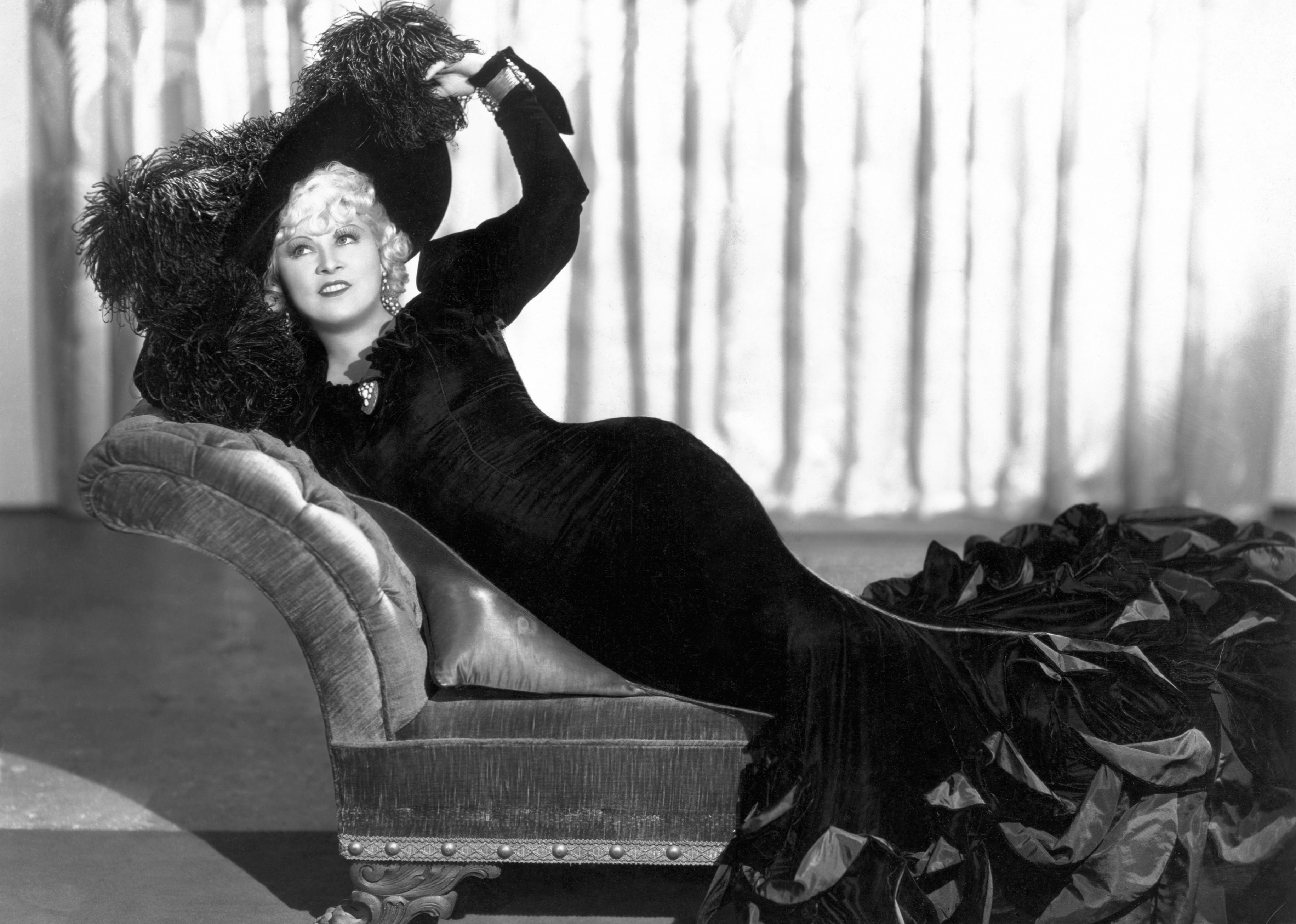 Mae West lying on a chaise lounge in a velvet gown and hat.