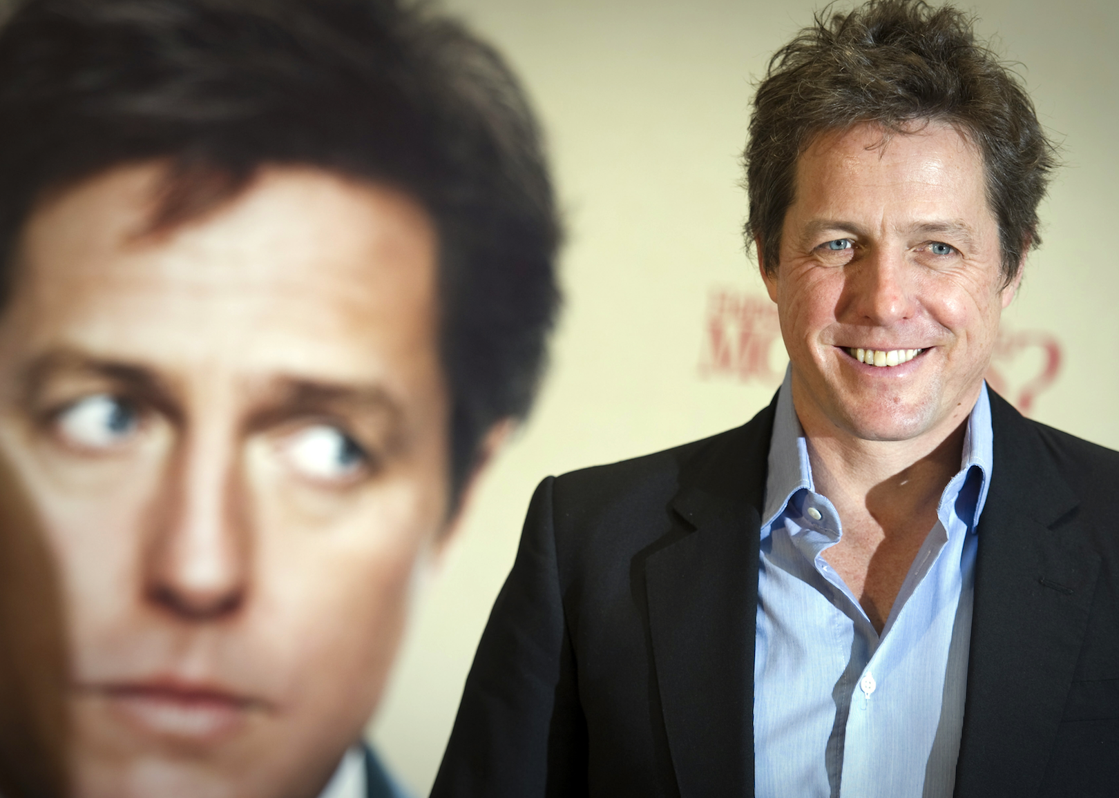 Hugh Grant smiling in front of a background poster of himself.