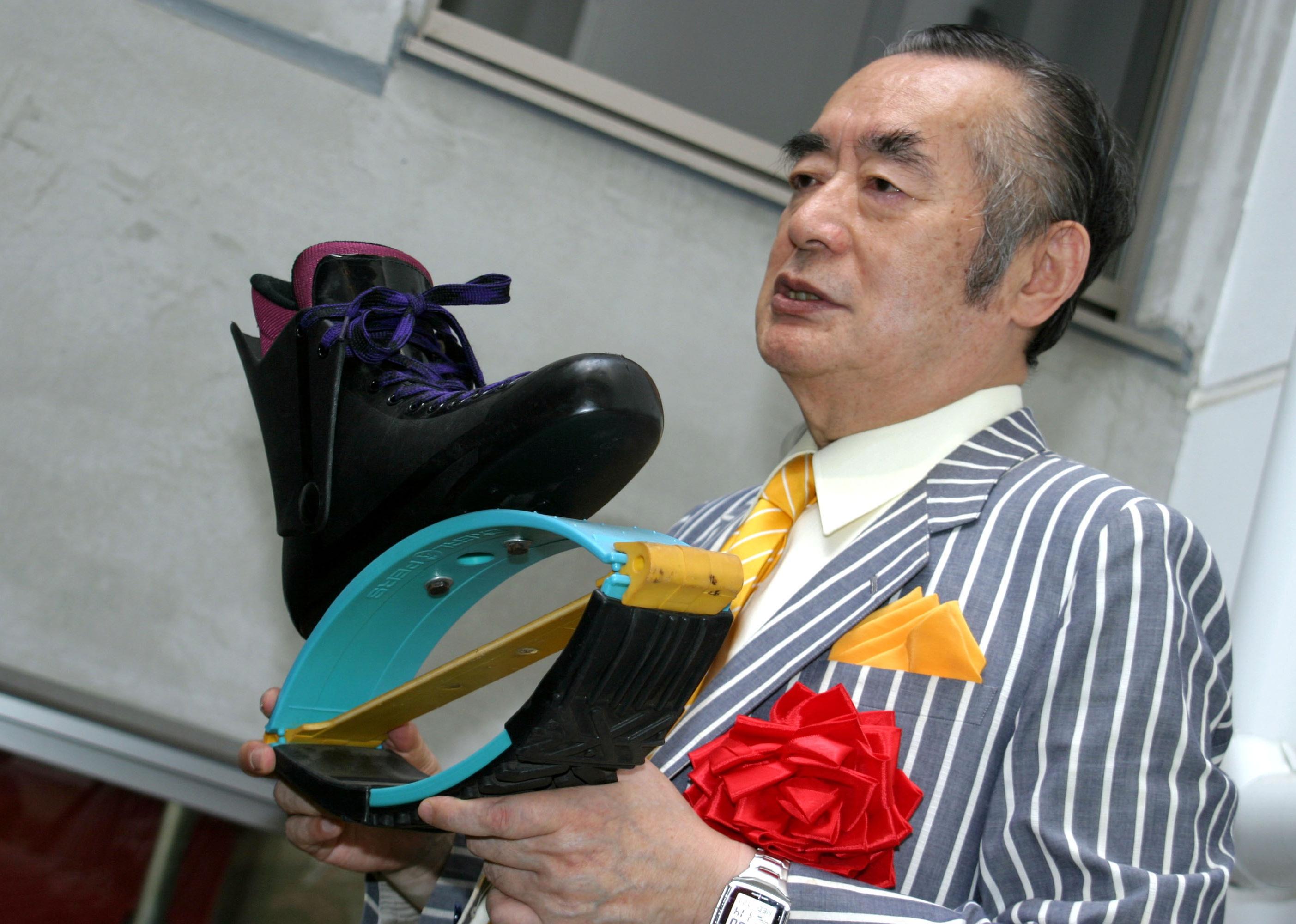 Yoshiro Nakamatsu, wearing a gray pinstripe suit with a yellow tie and red bow, holding a shoe invention.