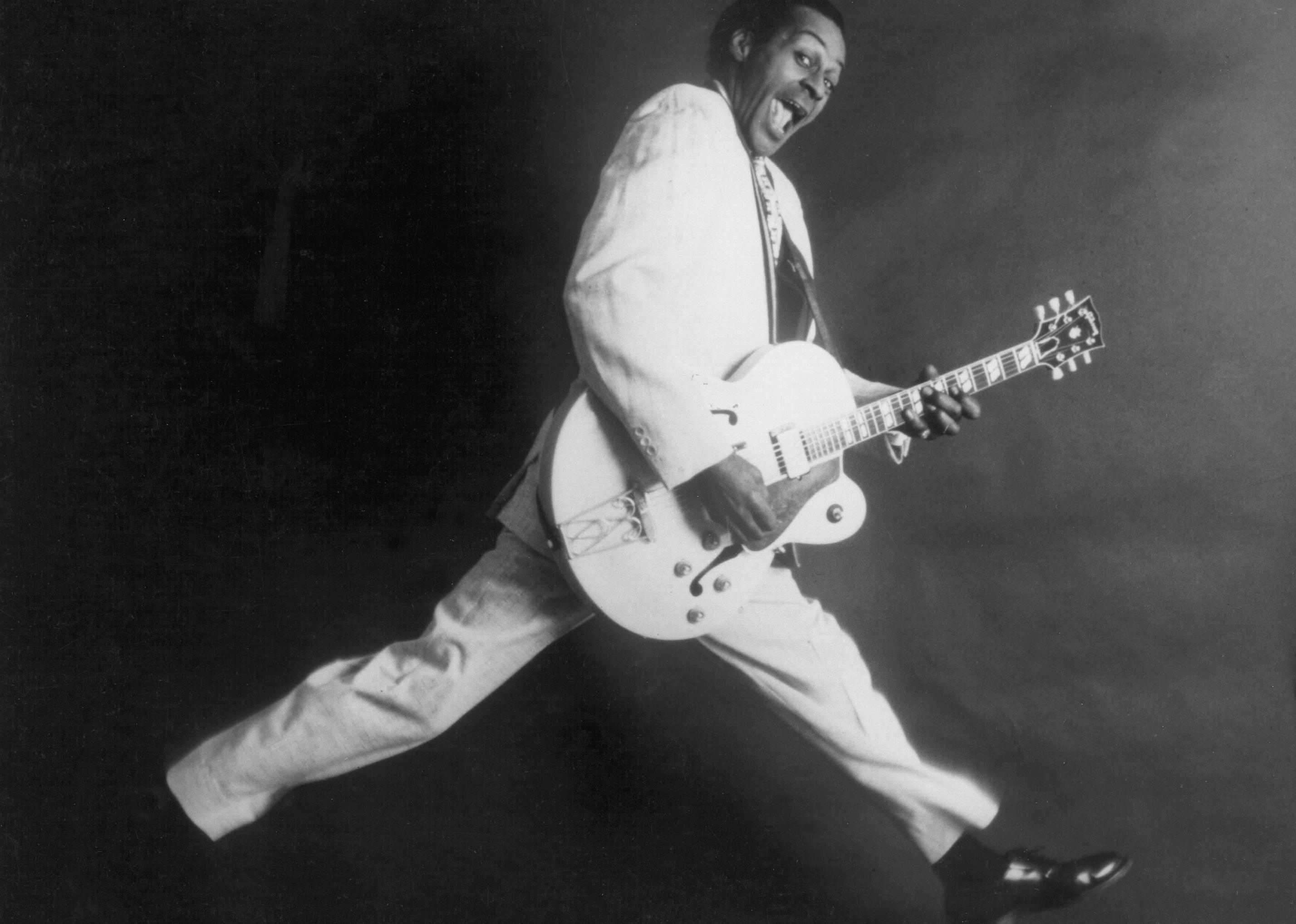Chuck Berry dancing with a guitar in a white suit.