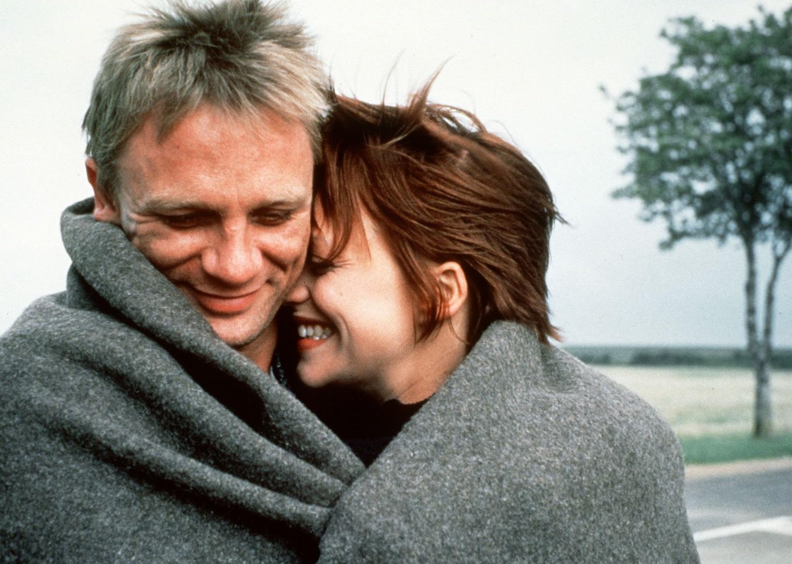 Daniel Craig and a woman with short red hair snuggling outside in a gray blanket.
