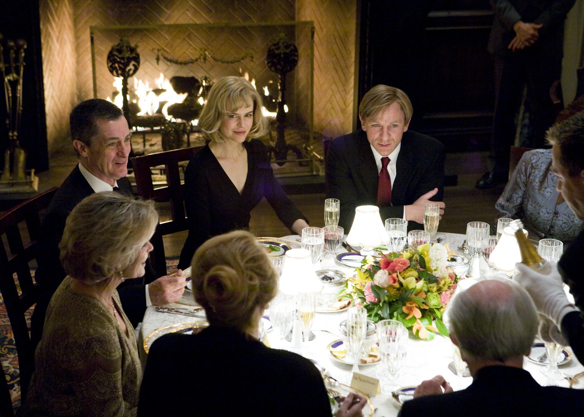 Nicole Kidman and Daniel Craig at a dining table set with flowers and fine china with a group in front of a fireplace.