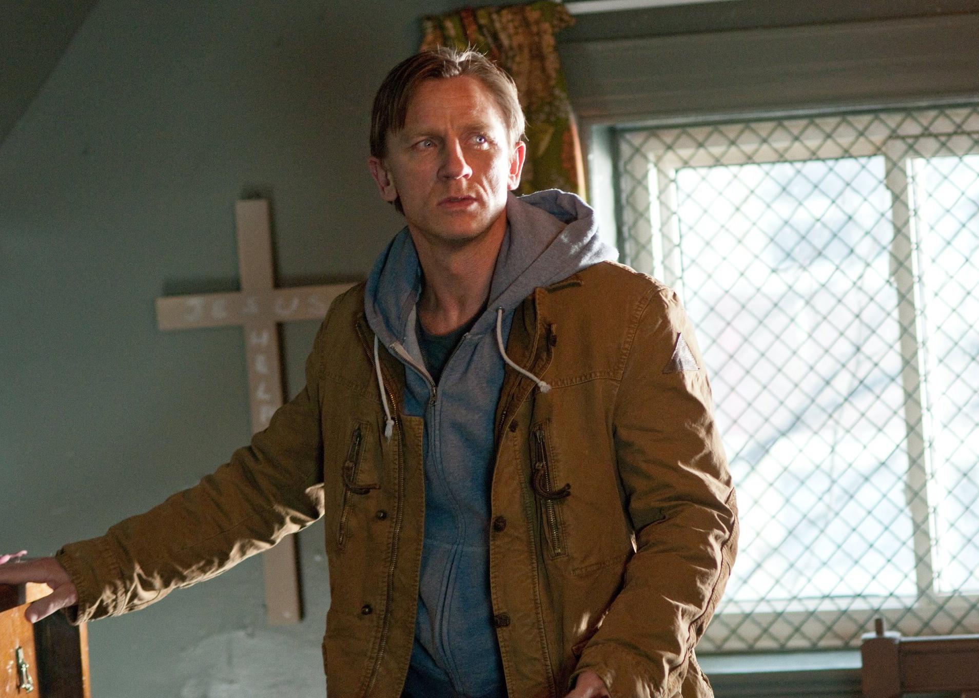 Daniel Craig in a hoodie and jacket in front of a cross.