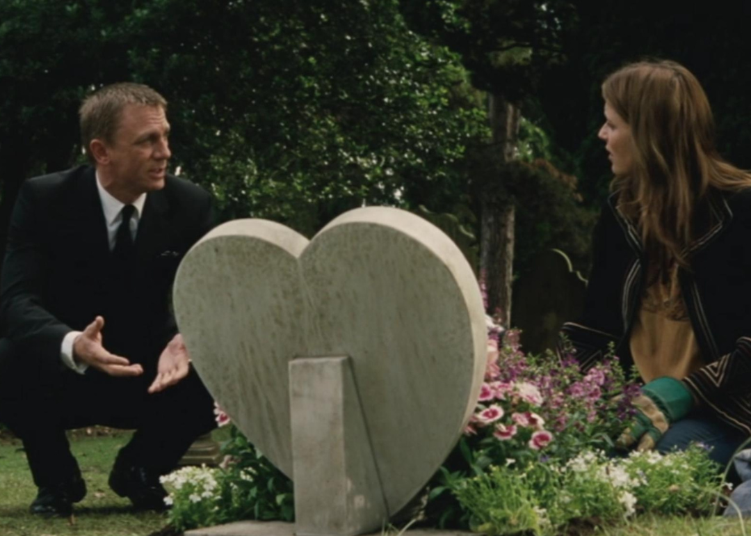 Daniel Craig talking with a woman on the ground in front of a heart-shaped gravestone covered in flowers.
