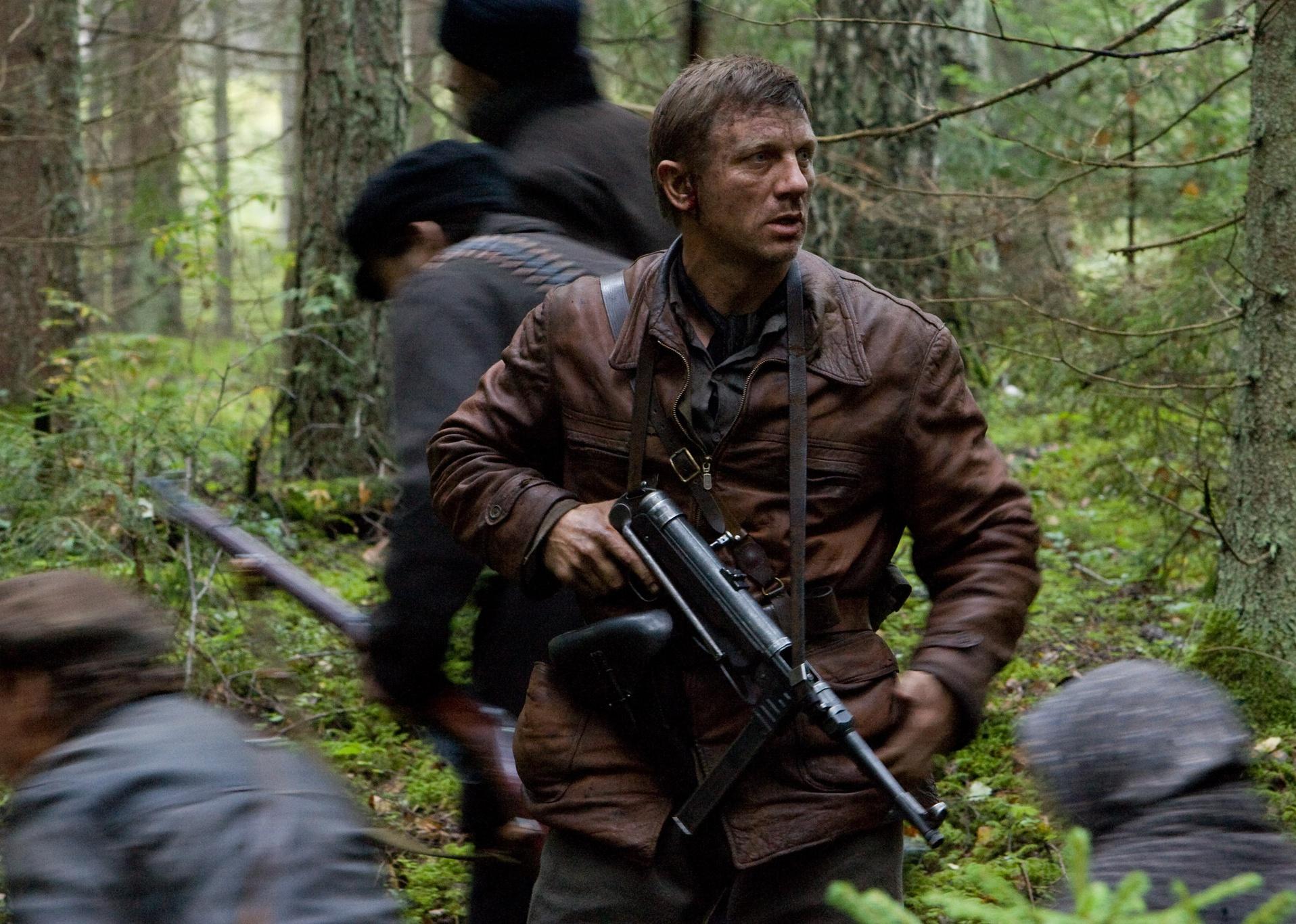 Daniel Craig in a brown leather jacket standing guard with a large gun around his chest as people pass through the woods.