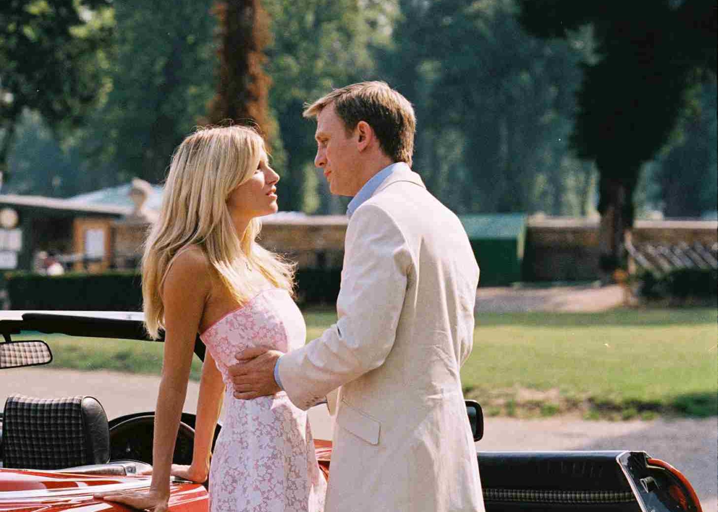 Daniel Craig in a white suit leaning over to kiss Sienna Miller next to a red classic convertible car.