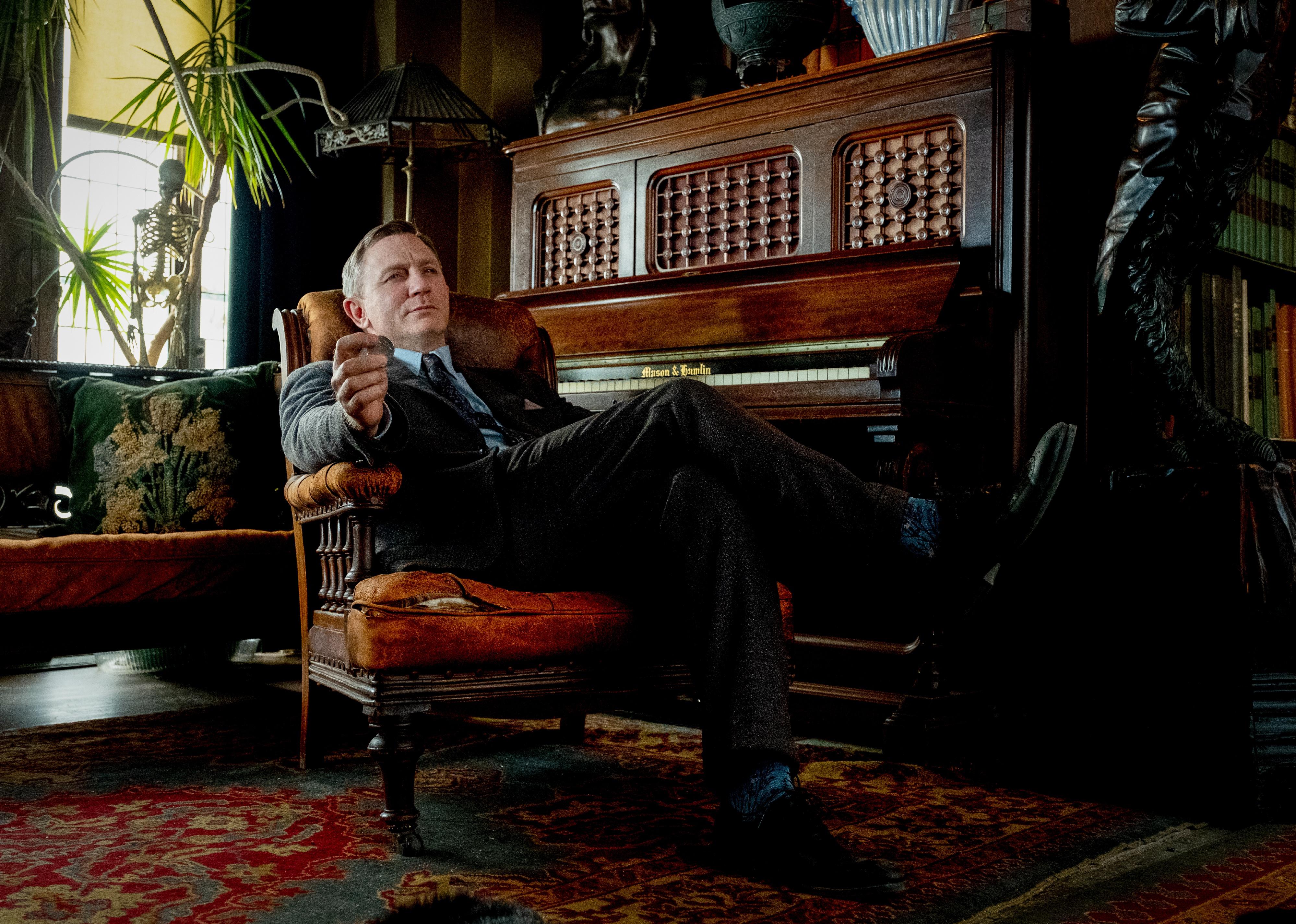 Daniel Craig in a suit sitting in a chair next to a piano in a dark wood room.