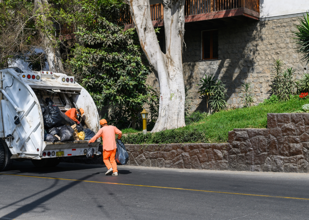 Two men in orange put garbage bags into a refuse truck.