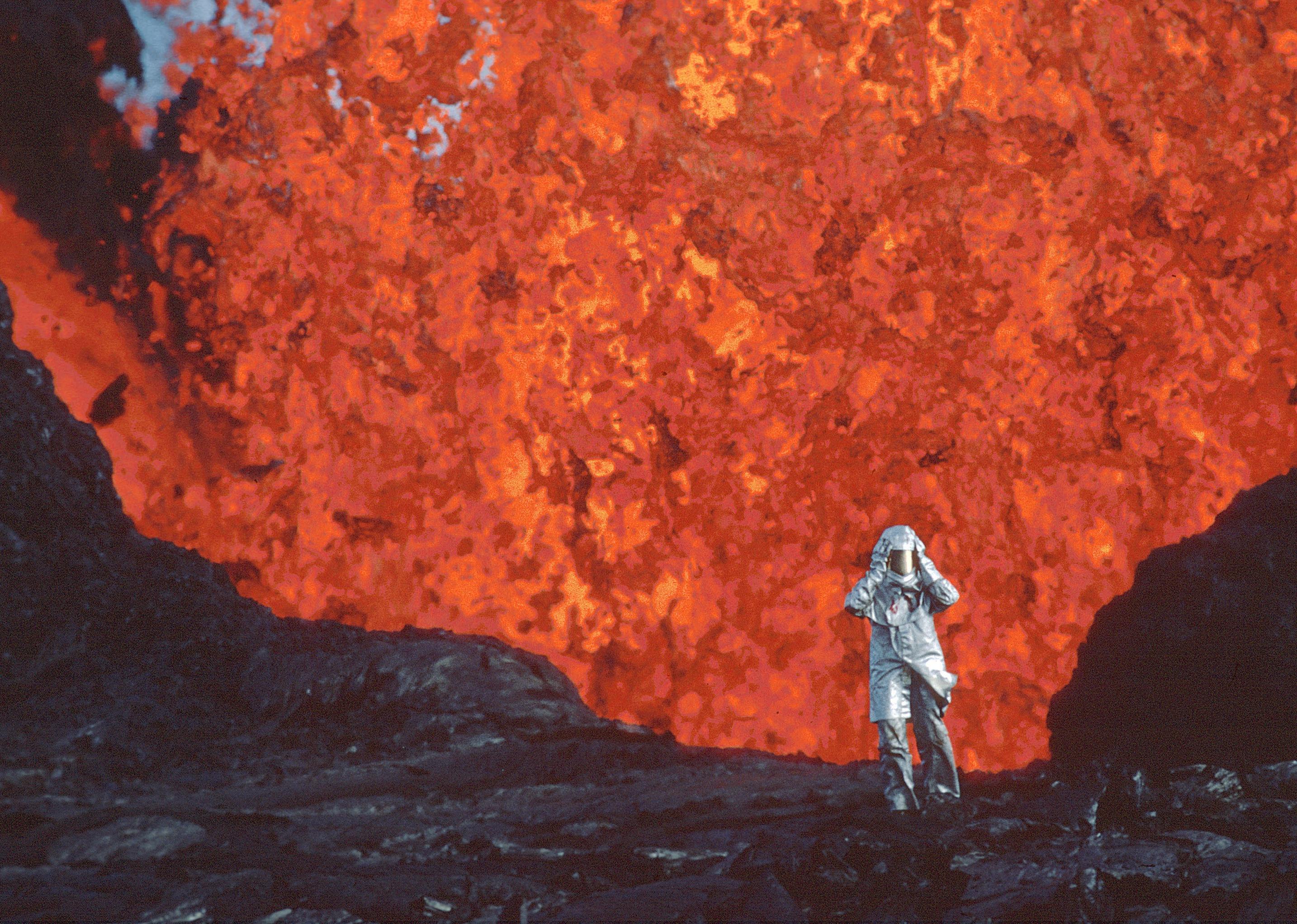 A person in a silver suit walking away from an erupting volcano.