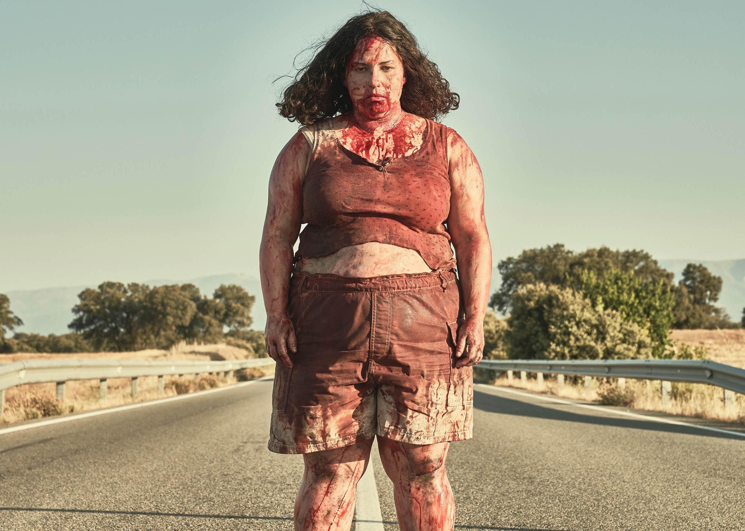 A woman standing on a highway in shorts and a tank top covered in blood.