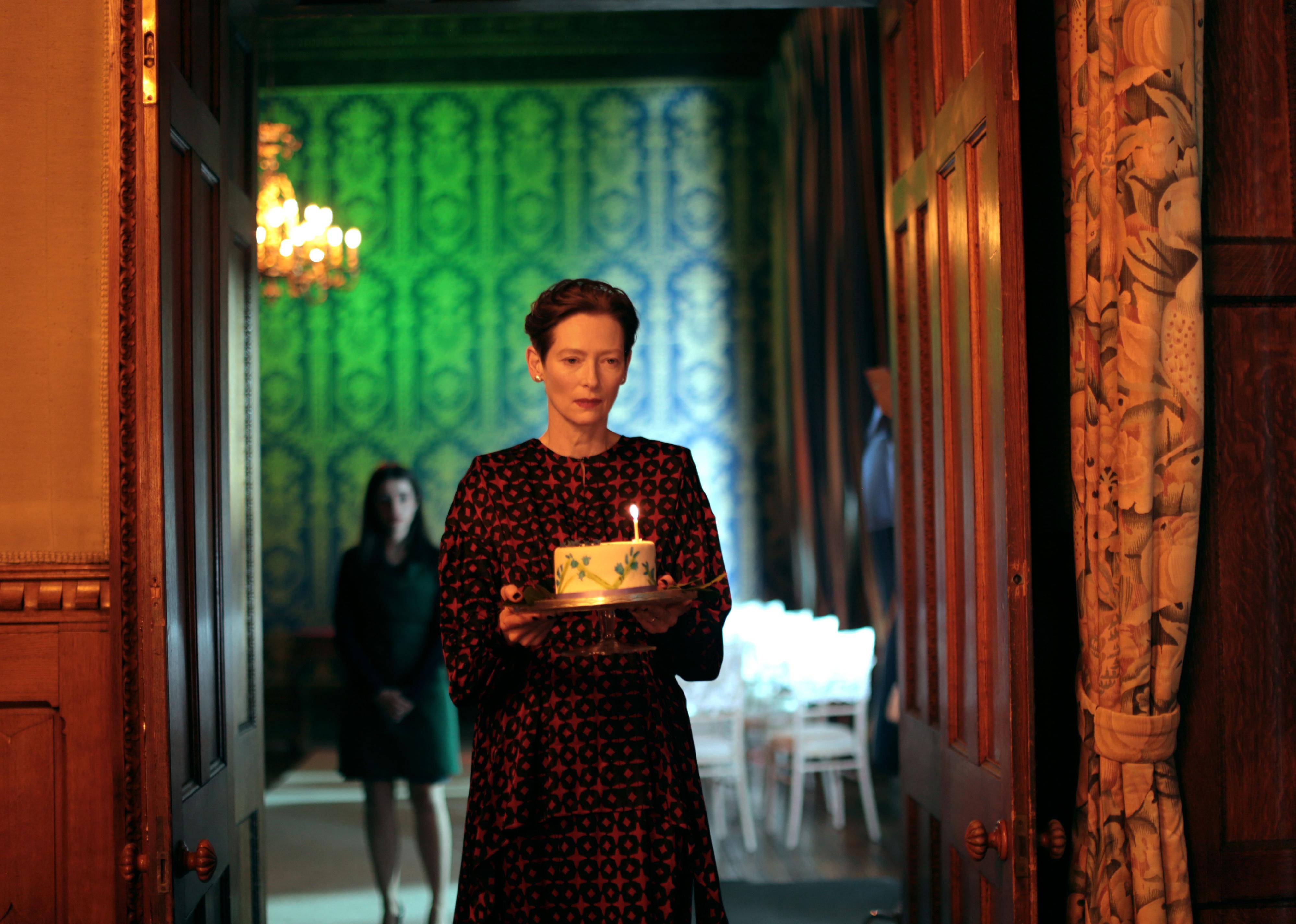 Tilda Swinton carrying a cake with one lit candle down an ornate historic looking hallway.