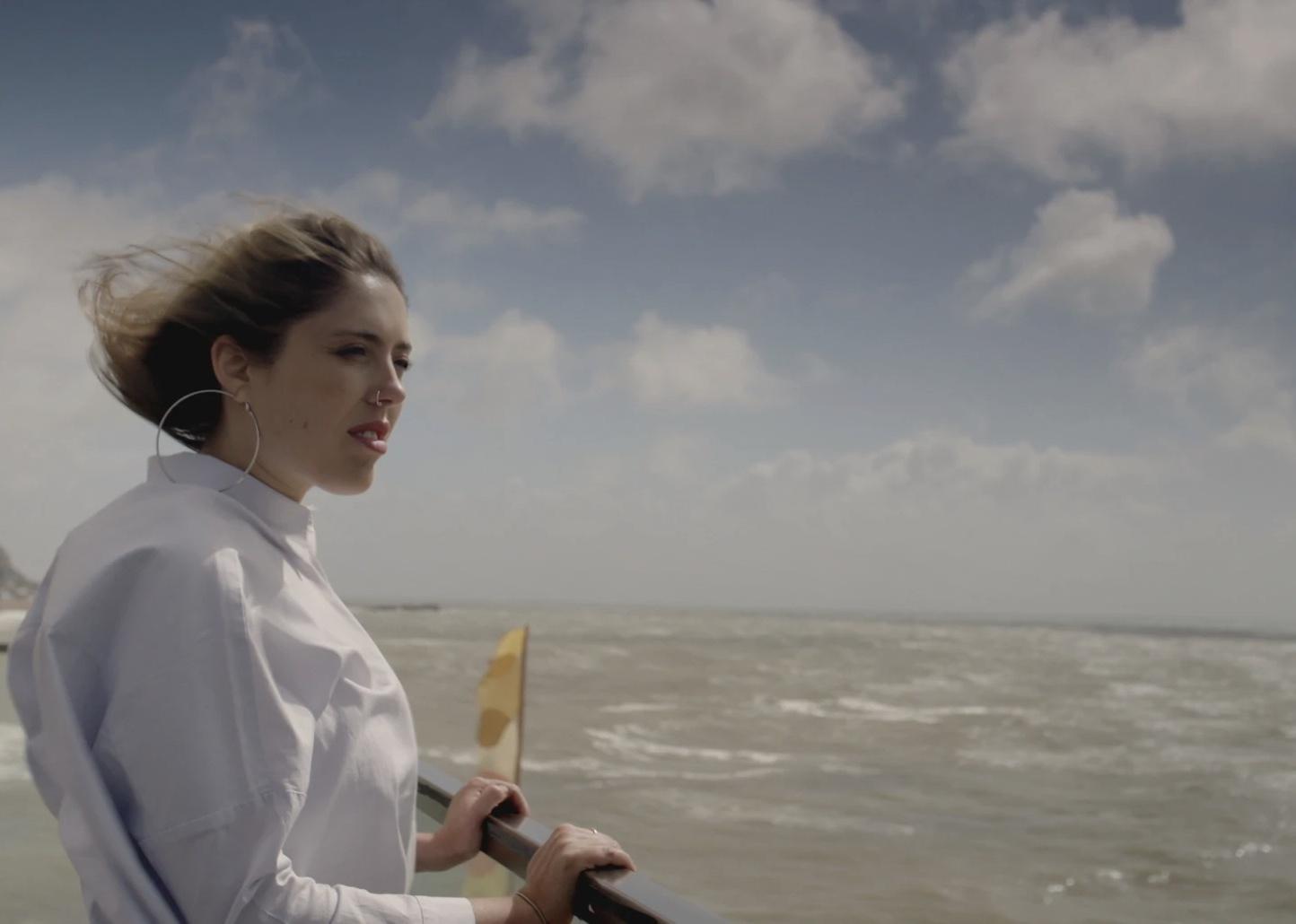 A woman in white with a nose ring and large hoop earrings stands next to a windy sea.