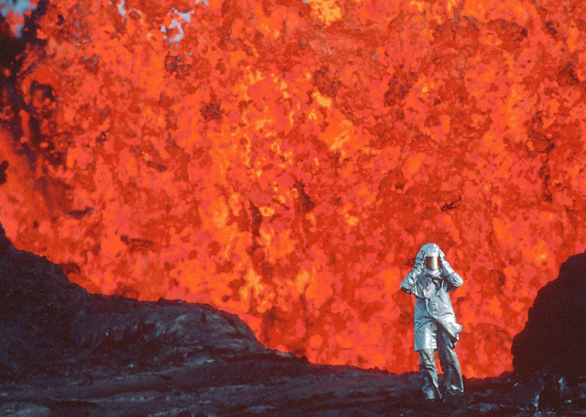 A person walking from a spewing volcano in a silver suit and helmet.