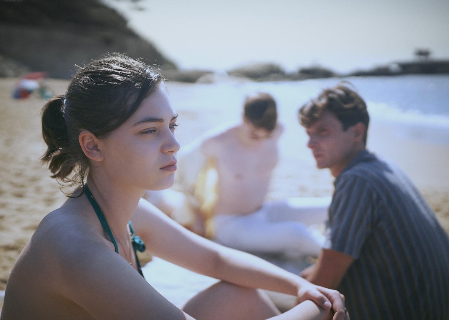 A girl sitting on the beach looking out at the ocean with two men in the background.