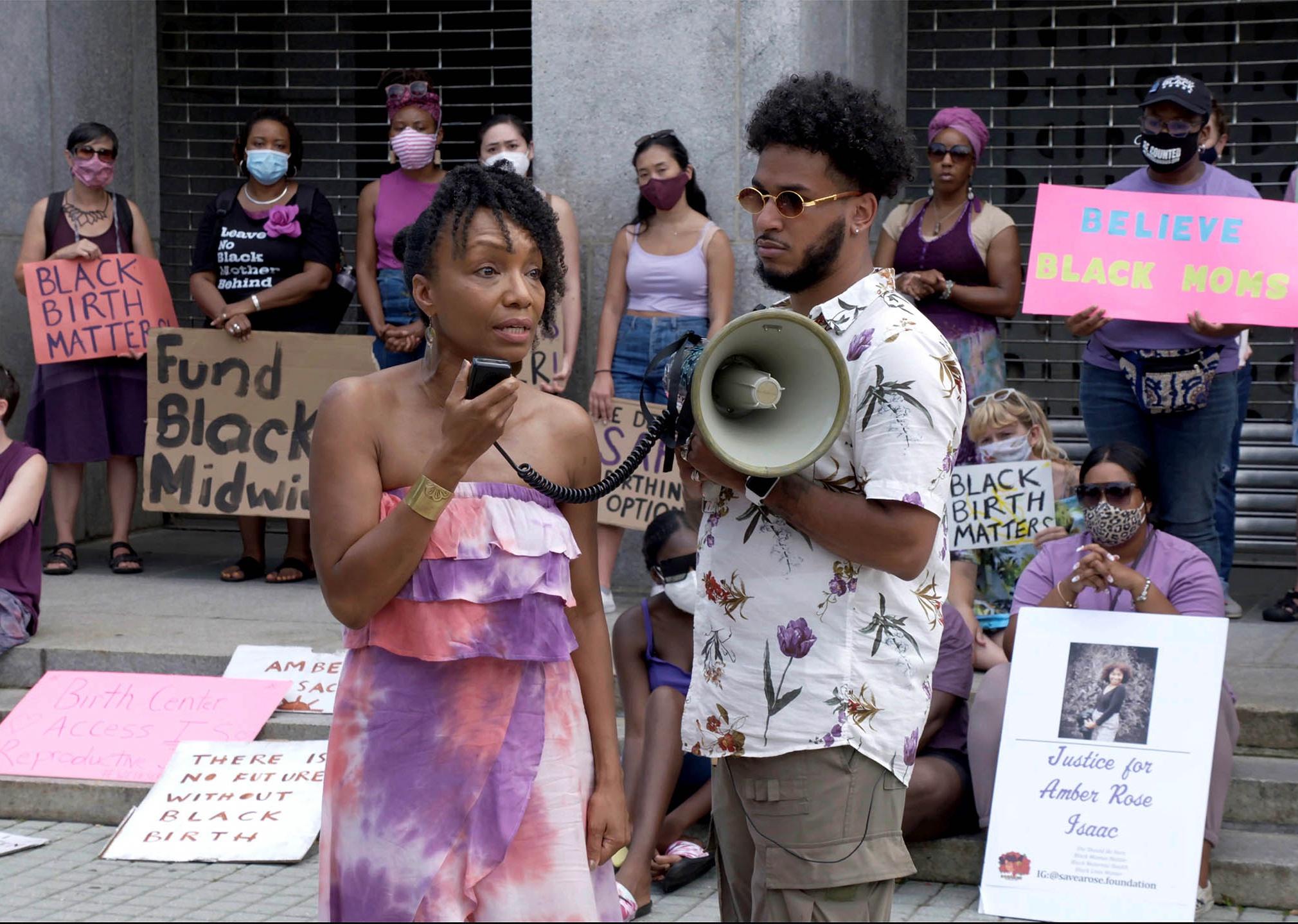 A man and woman holding a megaphone and speaking on steps surrounded by protesters.