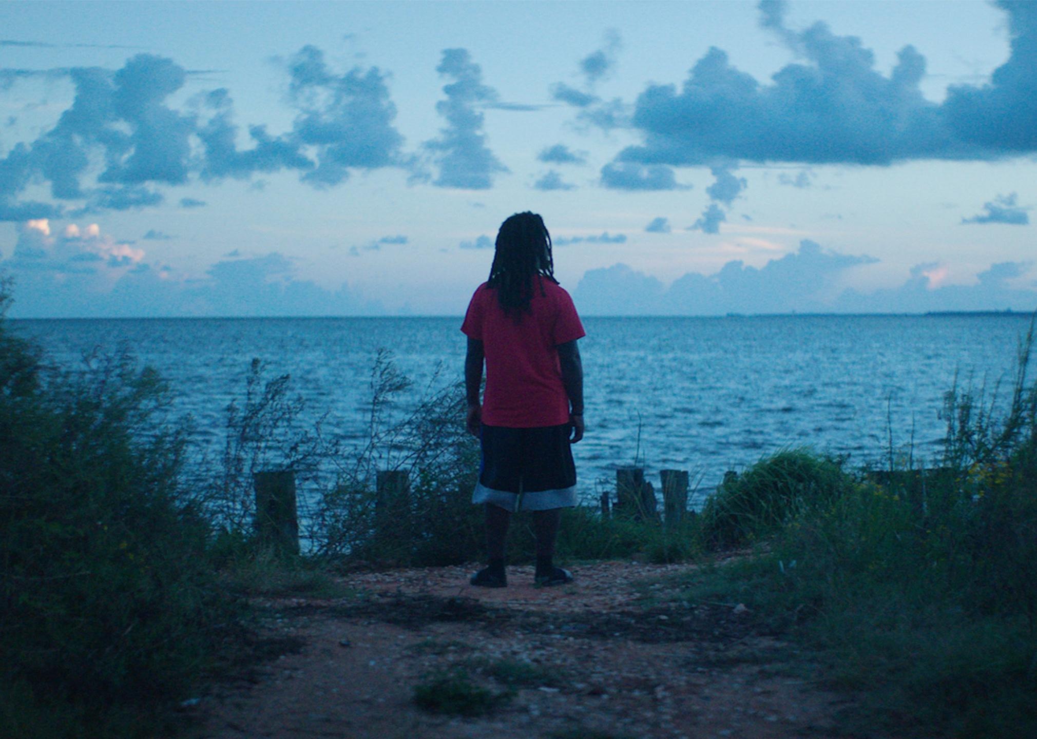 A black man standing at the shore looking out at the water.