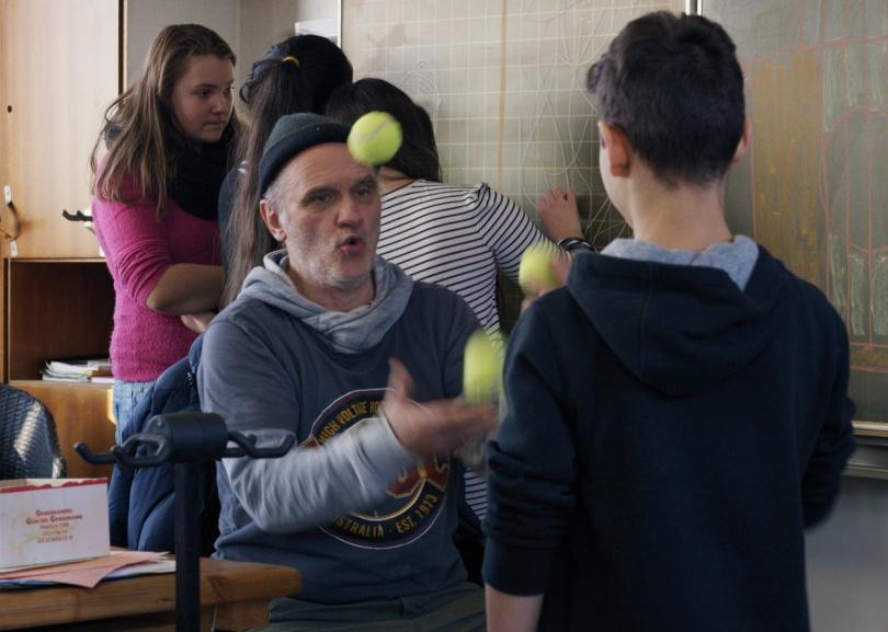A man in a beanie juggling tennis balls in a classroom.