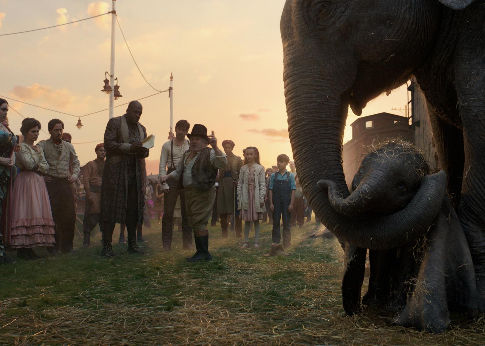Danny DeVito and Colin Farrell talking to a crowd of people outside a circus tent next to a baby and mother elephant.