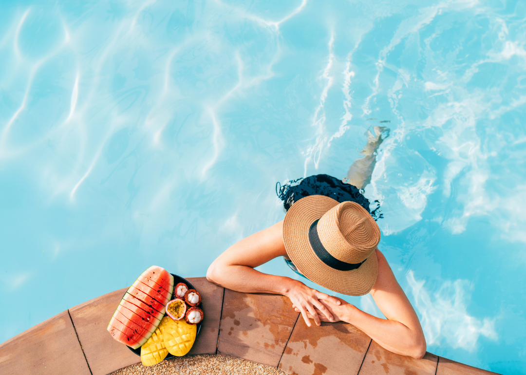 A person in the pool wearing a wide brimmed hat with fruit next to them.