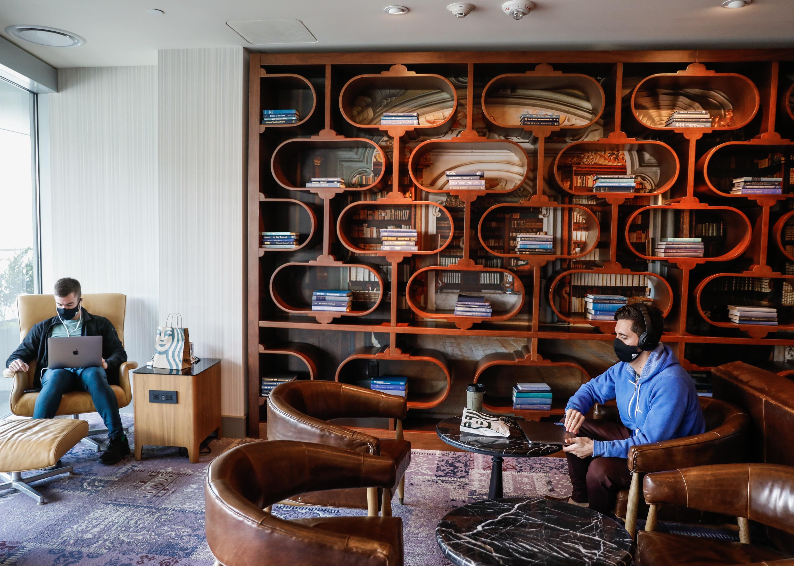 Two men working in a common work space with an artistic bookshelf on the wall.