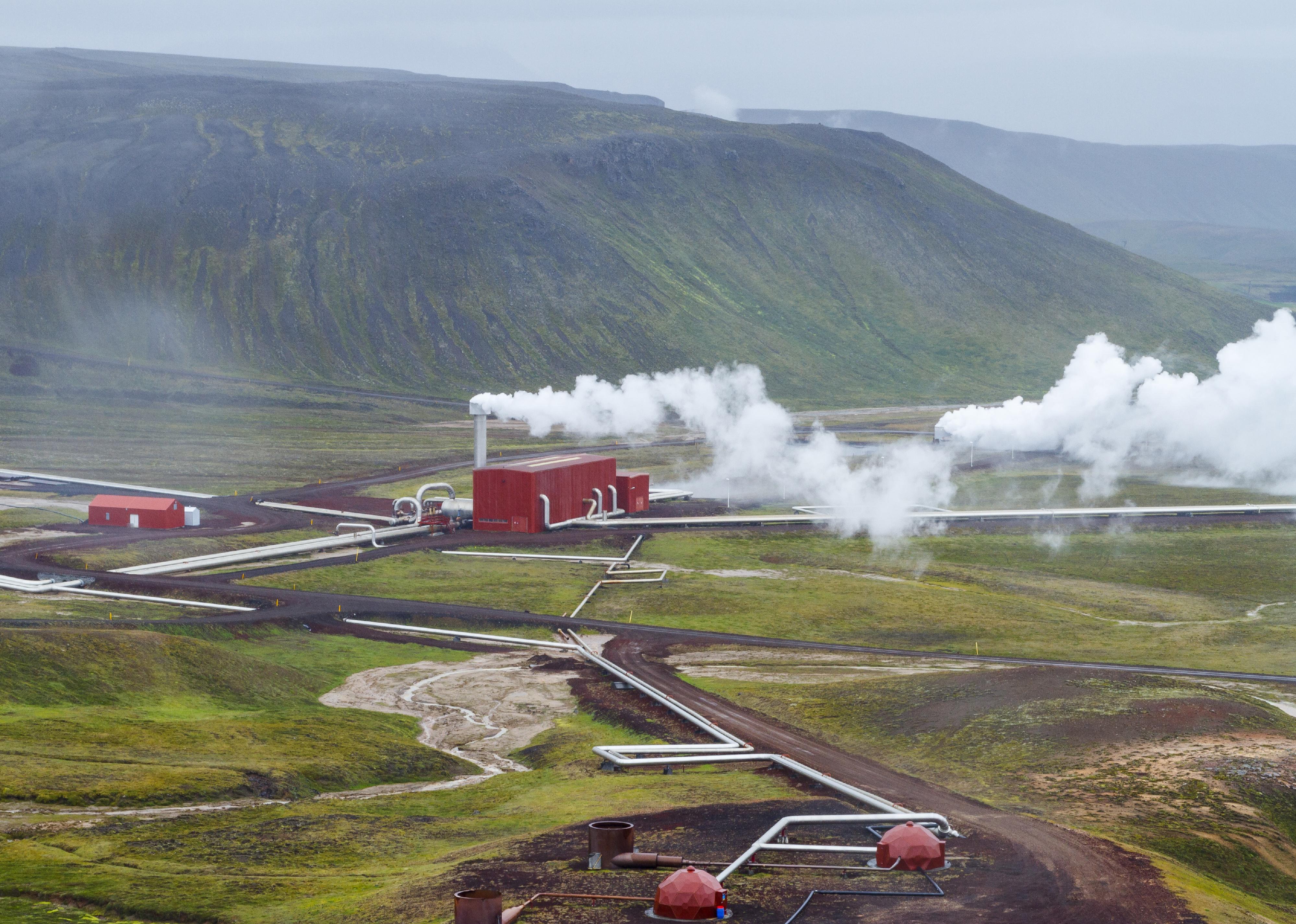 A series of red buildings and pipes at a geothermal power station surrounded by green land and mountains in Iceland.