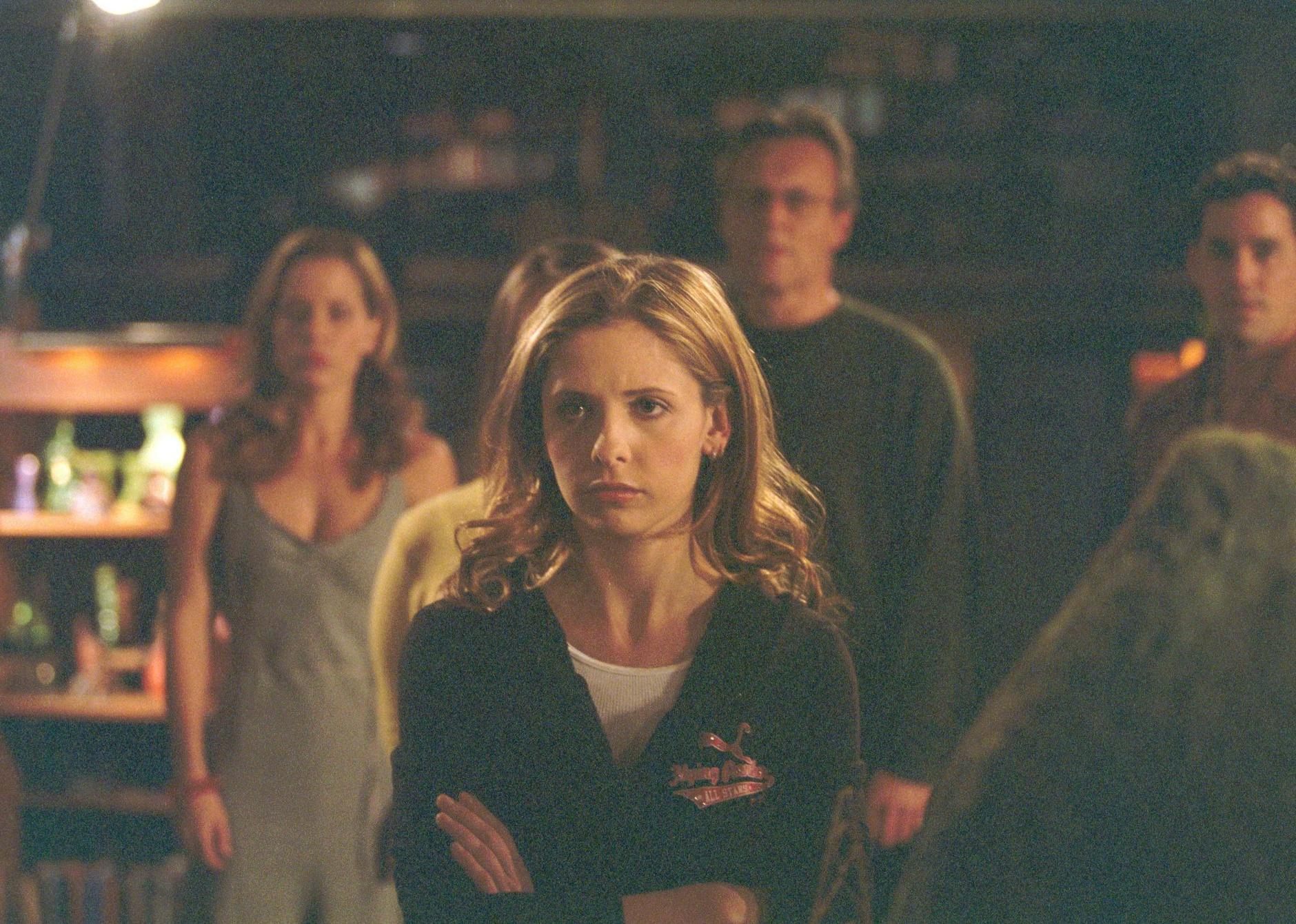 Sarah Michelle Gellar standing with arms crossed in front of a group of people.