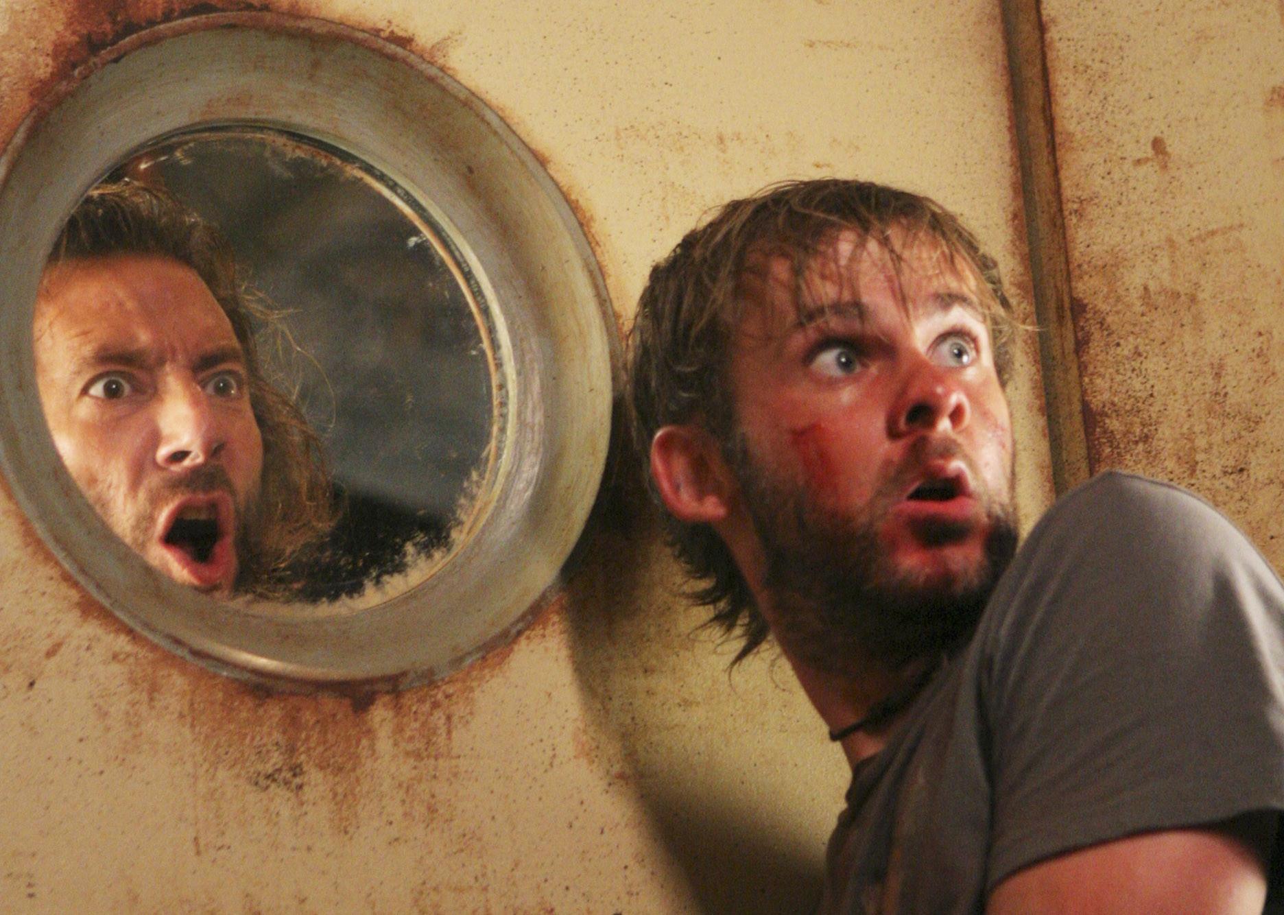 Two men stand looking shocked with a door and a small porthole window between them.