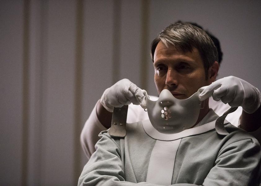 Mads Mikkelsen in a straighjacket with a mask being placed on his face.
