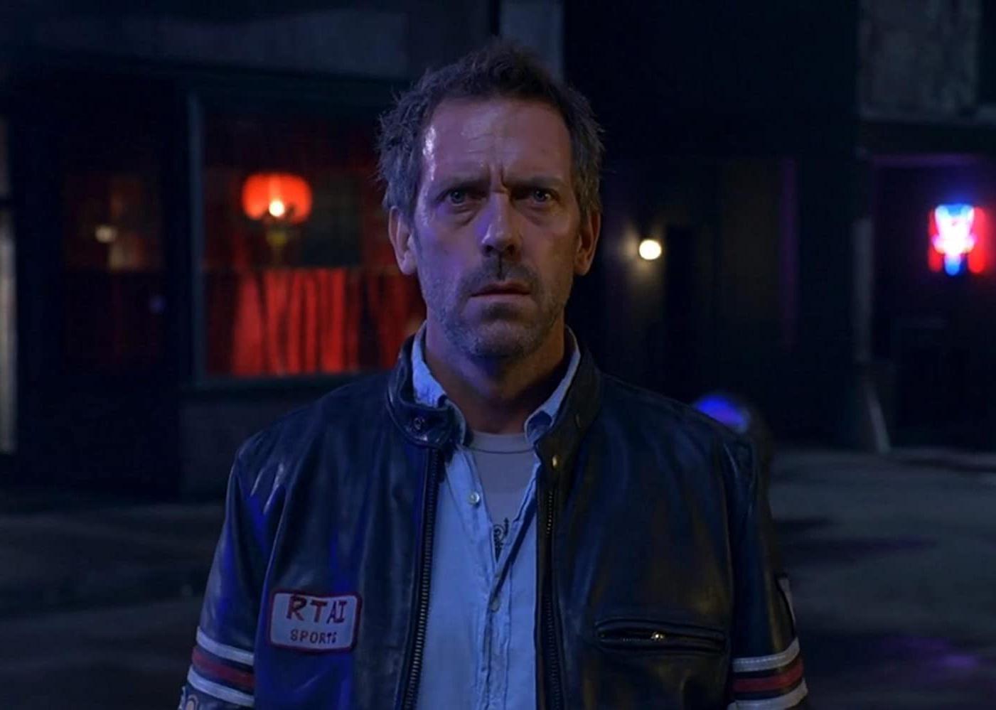 Hugh Laurie standing at a door in a black leather jacket.
