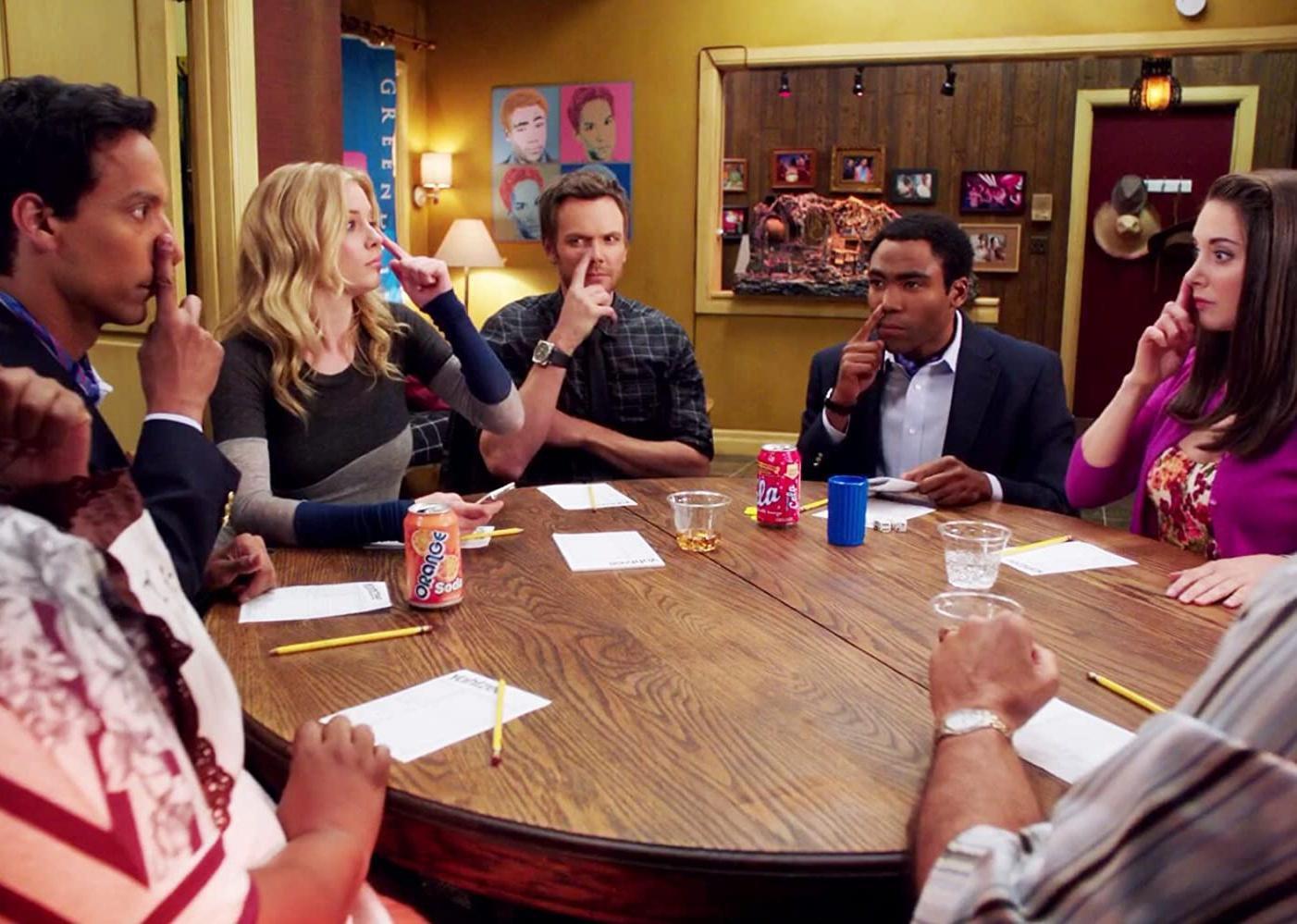 Joel McHale and a group of people sit around a table with their fingers on their noses.