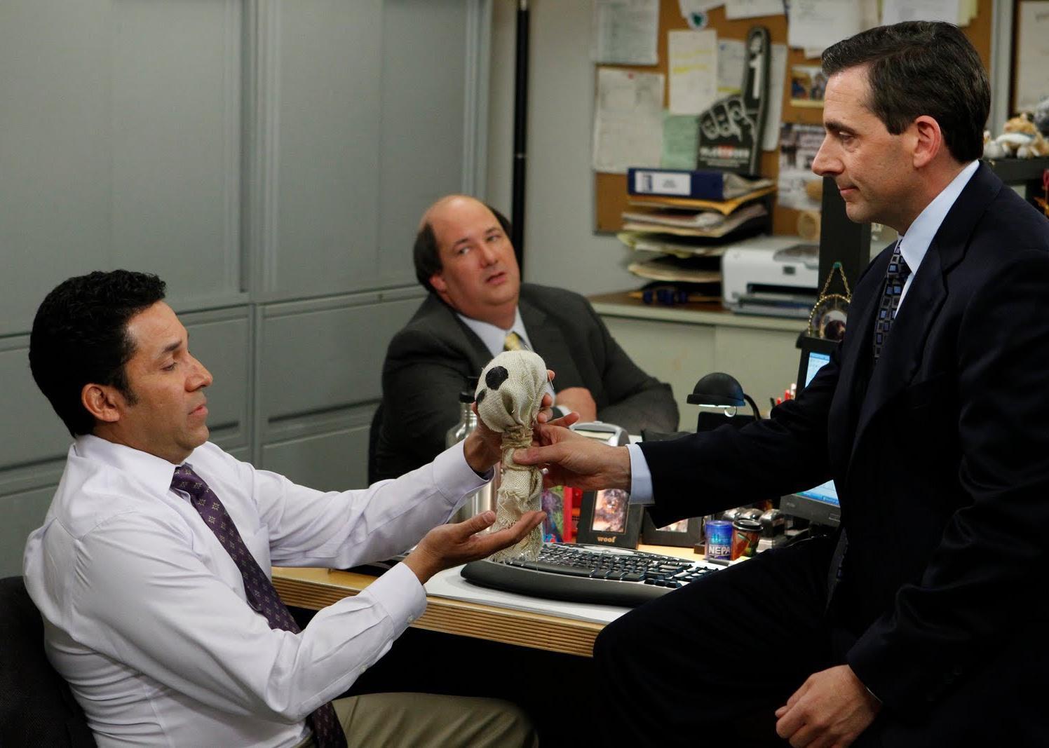 Steve Carell hands a burlap object to another man in an office.