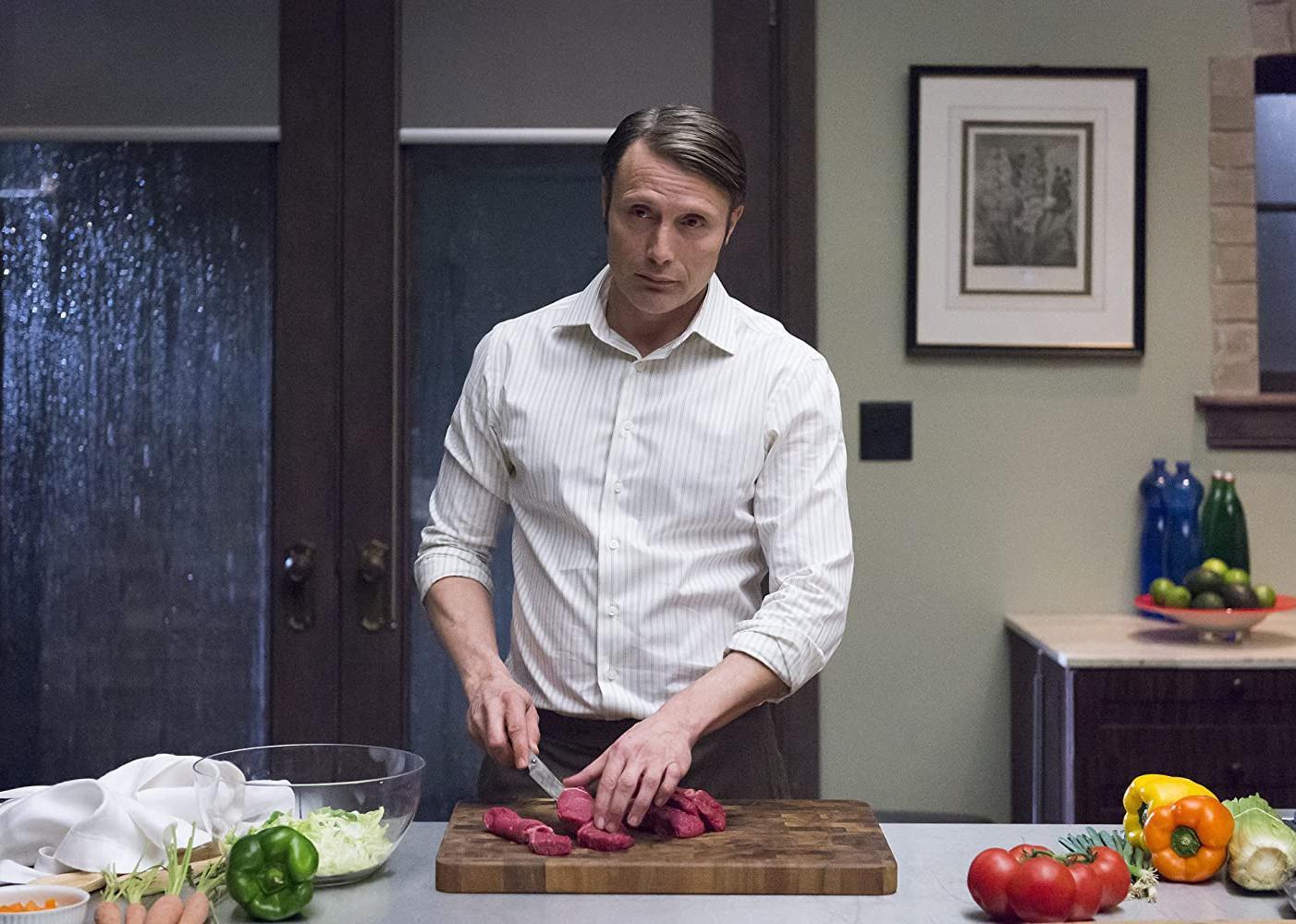 Mads Mikkelsen chops raw meat on a kitchen island covered in vegetables.