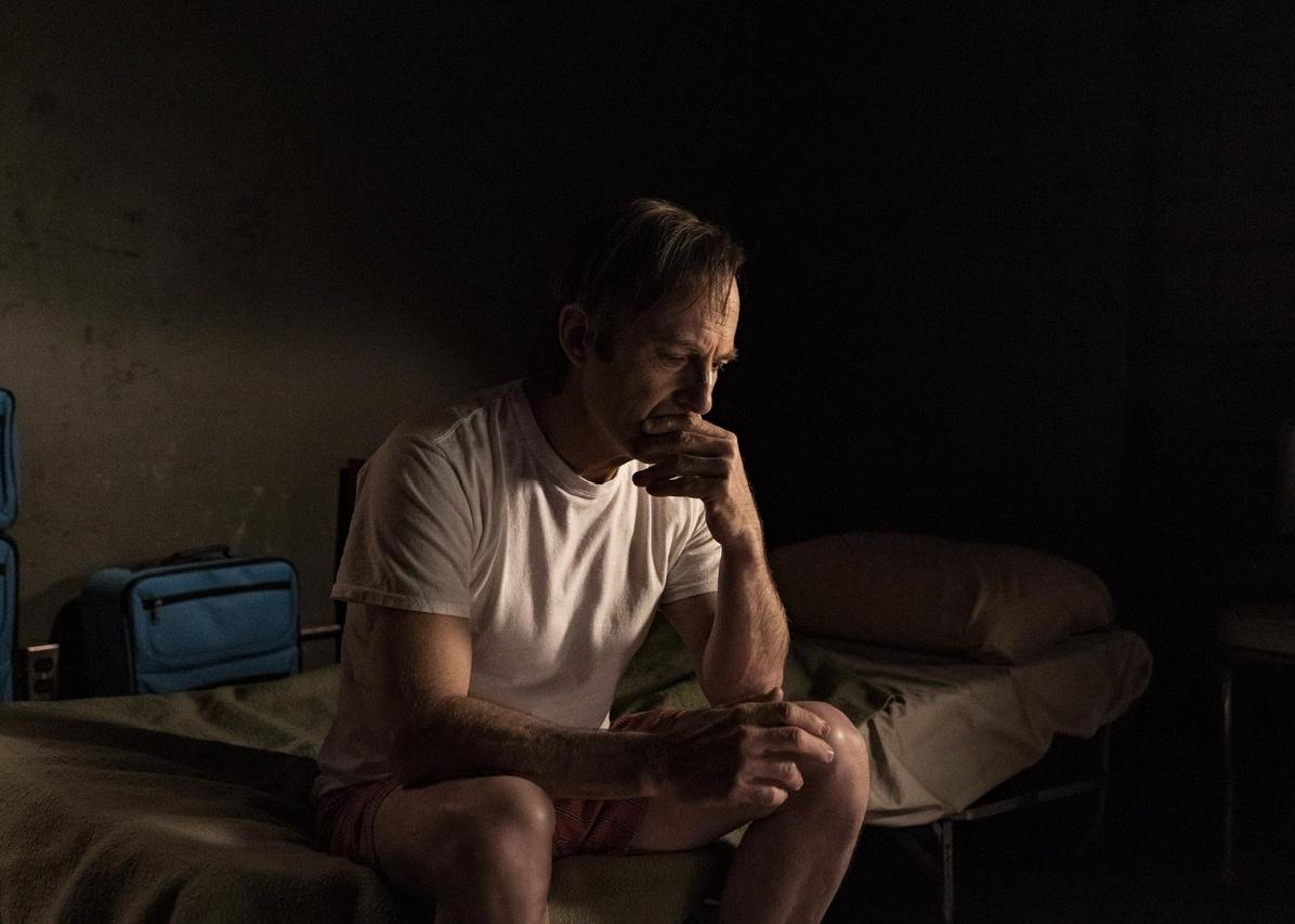 Bob Odenkirk in a t-shirt and shorts in a dark room sitting on a bed.