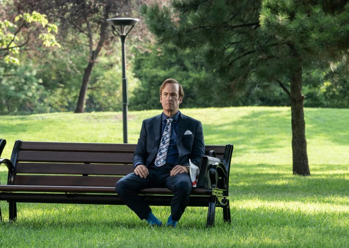 Bob Odenkirk sitting on a park bench with a briefcase and a package.