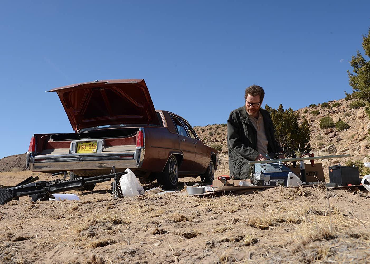 Bryan Cranston kneeling next to a car with a open truck surrounded by guns and tools.