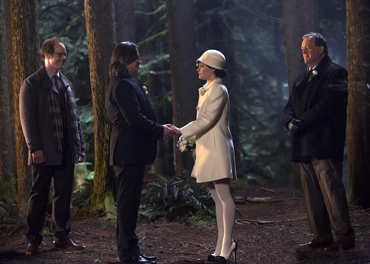 A man in a black suit and a woman in a white dress, coat and hat stand together in the woods holding hands with two others watching them.