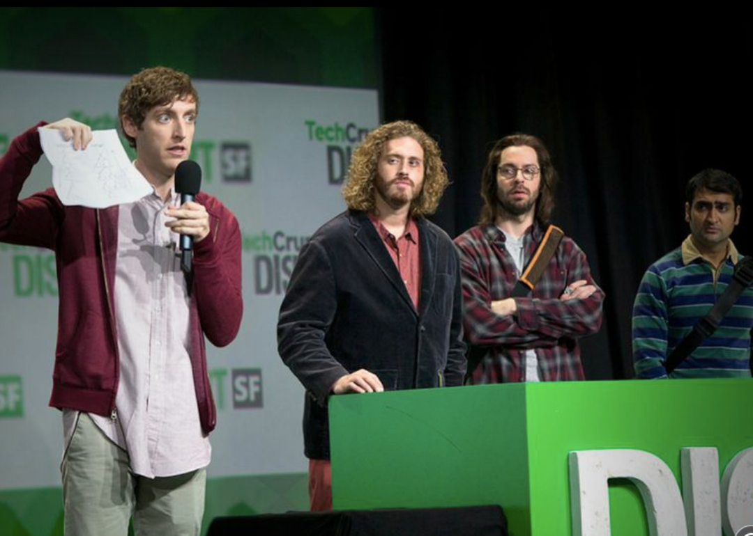 Four worried looking men stand on a stage, one presenting with a piece of paper.