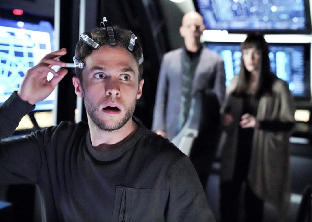 A man with clamps on his head in a room full of screens with two people behind him.