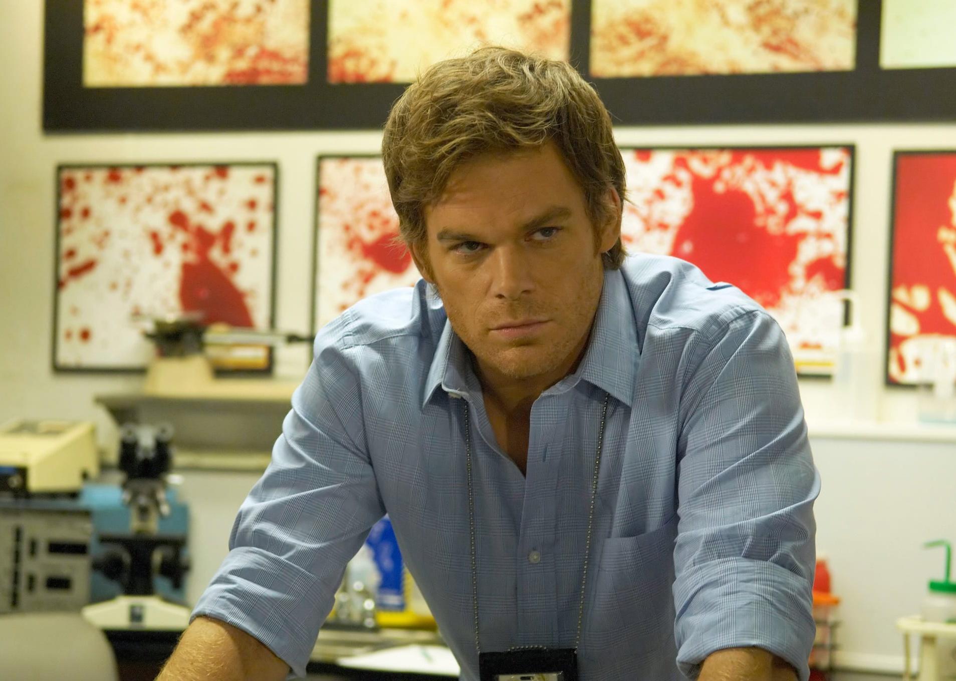 Michael C. Hall standing in an office wearing a badge in front of a wall of blood spatter images.