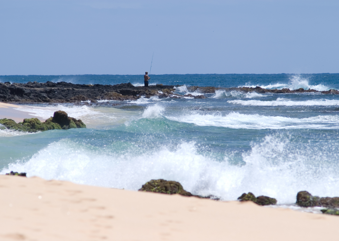 Waves crash onto the rocky, sandy shore while a person fishes in the distance. 