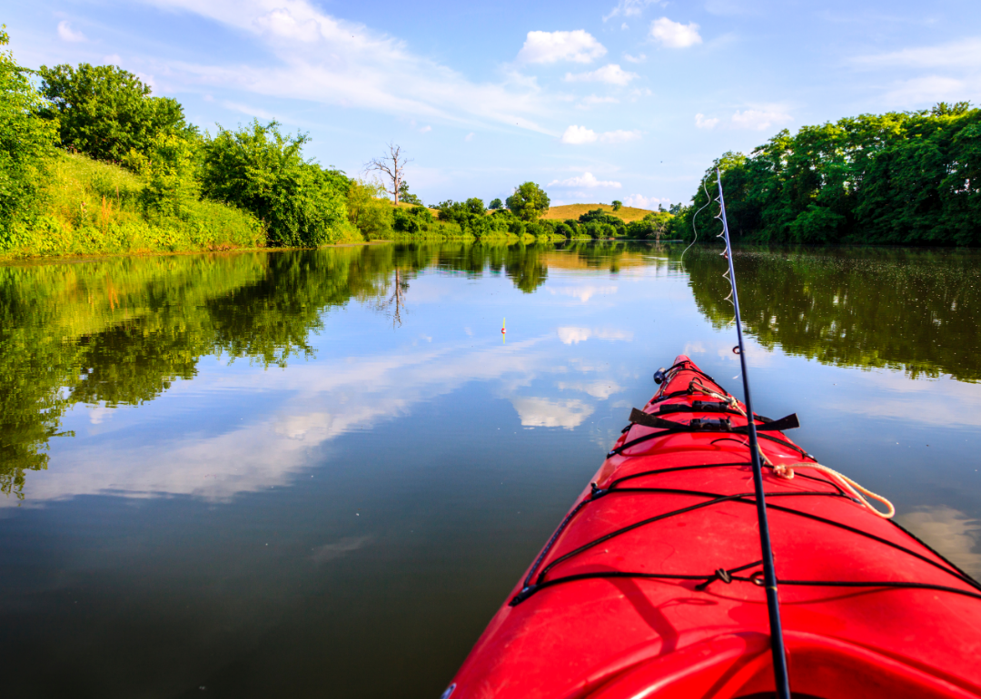 A red kayak with a fishing pole makes its way down a calm river lined with trees. 