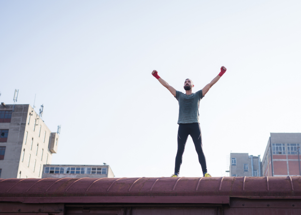 Male athlete standing on top of a building with arms stretched toward the sky looking satisfied.