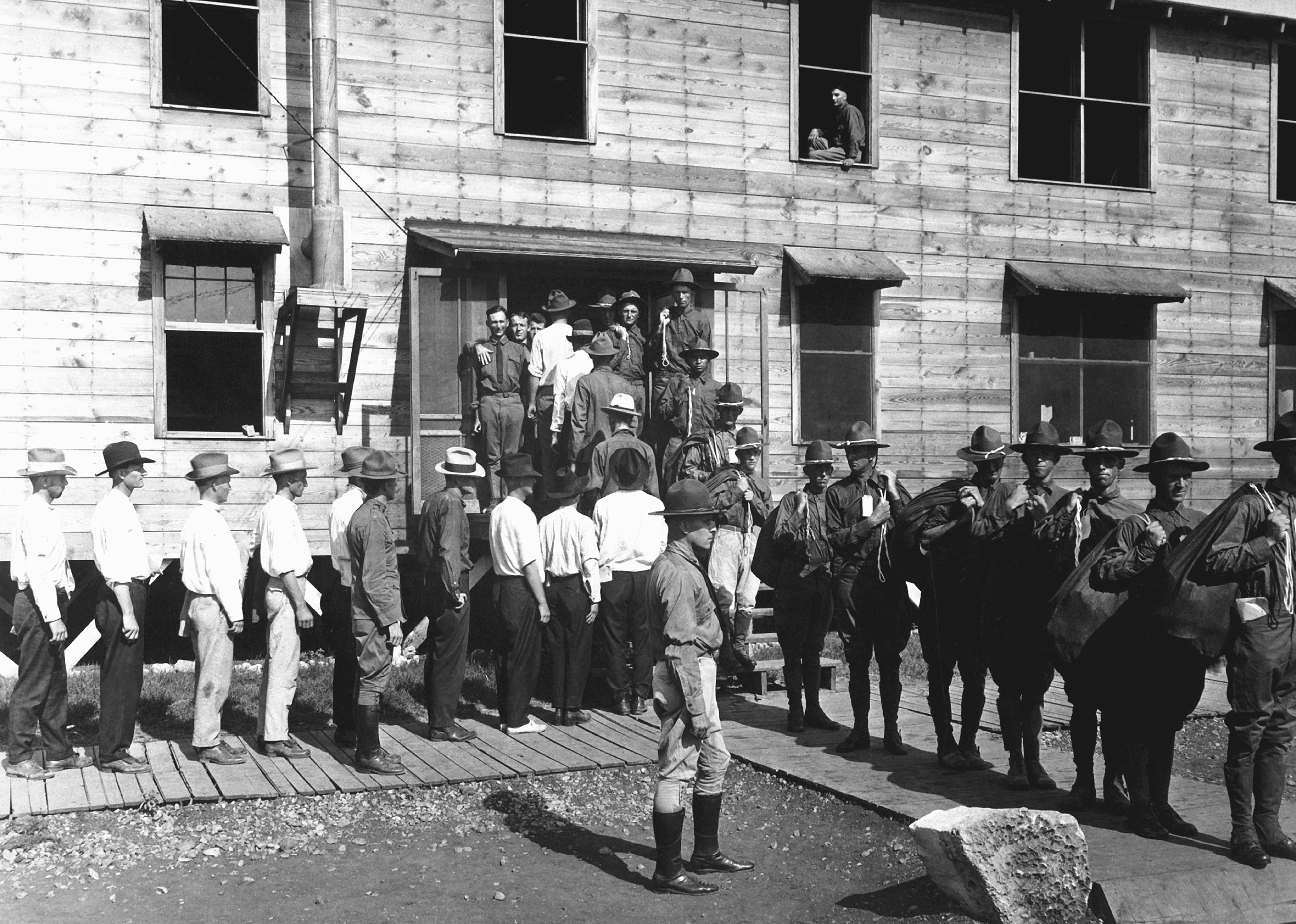 A line of civilian men, drafted for service, entering a building to report for duty and exiting in uniform.